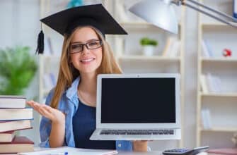 Молодая женщина в выпускной кепке и очках показывает ноутбук с пустым экраном в окружении книг за учебным столом.