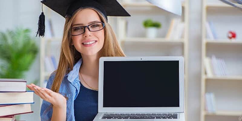 Молодая женщина в выпускной кепке и очках показывает ноутбук с пустым экраном в окружении книг за учебным столом.